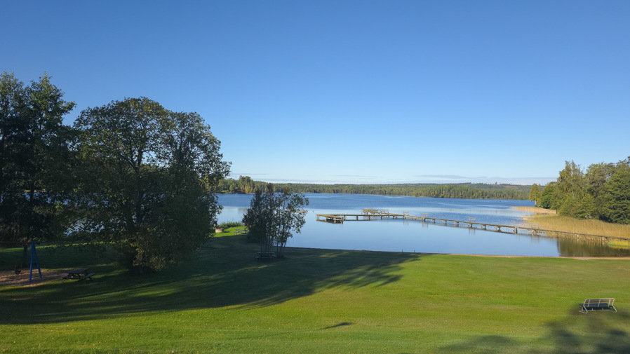 Utsikt över en sjö och en badplats med en brygga. Sjön är omringad utav träd.