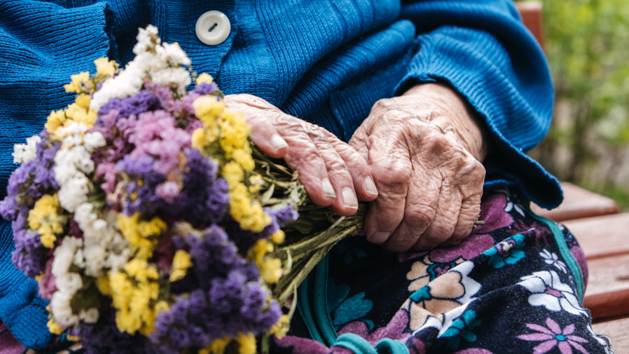 En äldre kvinna sitter på en bänk och håller i en blombukett.