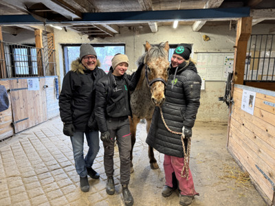 Tre personer och en h&auml;st st&aring;r i ett stall.