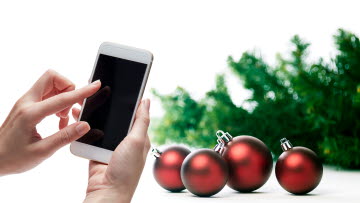 Hand håller i mobiltelefon, i bakgrunden julgran och julgranskulor